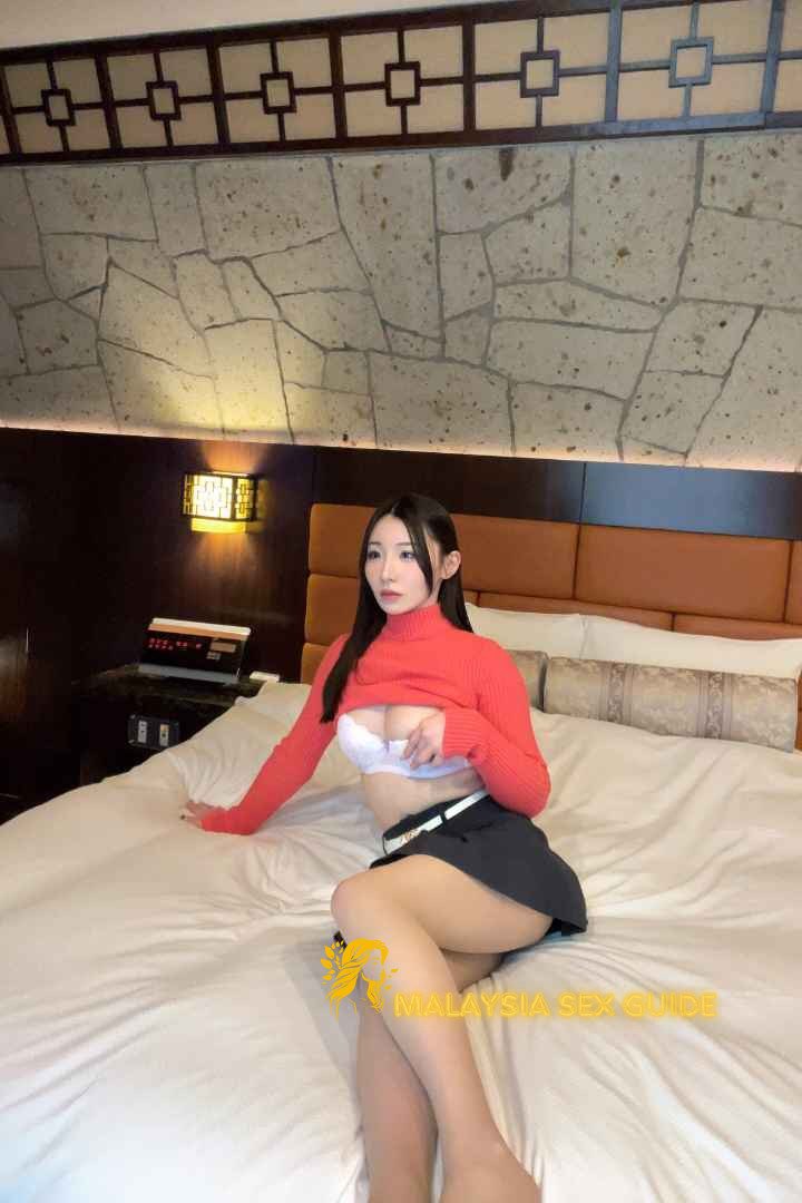 Attractive woman in red sweater and short skirt on a hotel bed. Malaysia escort services.